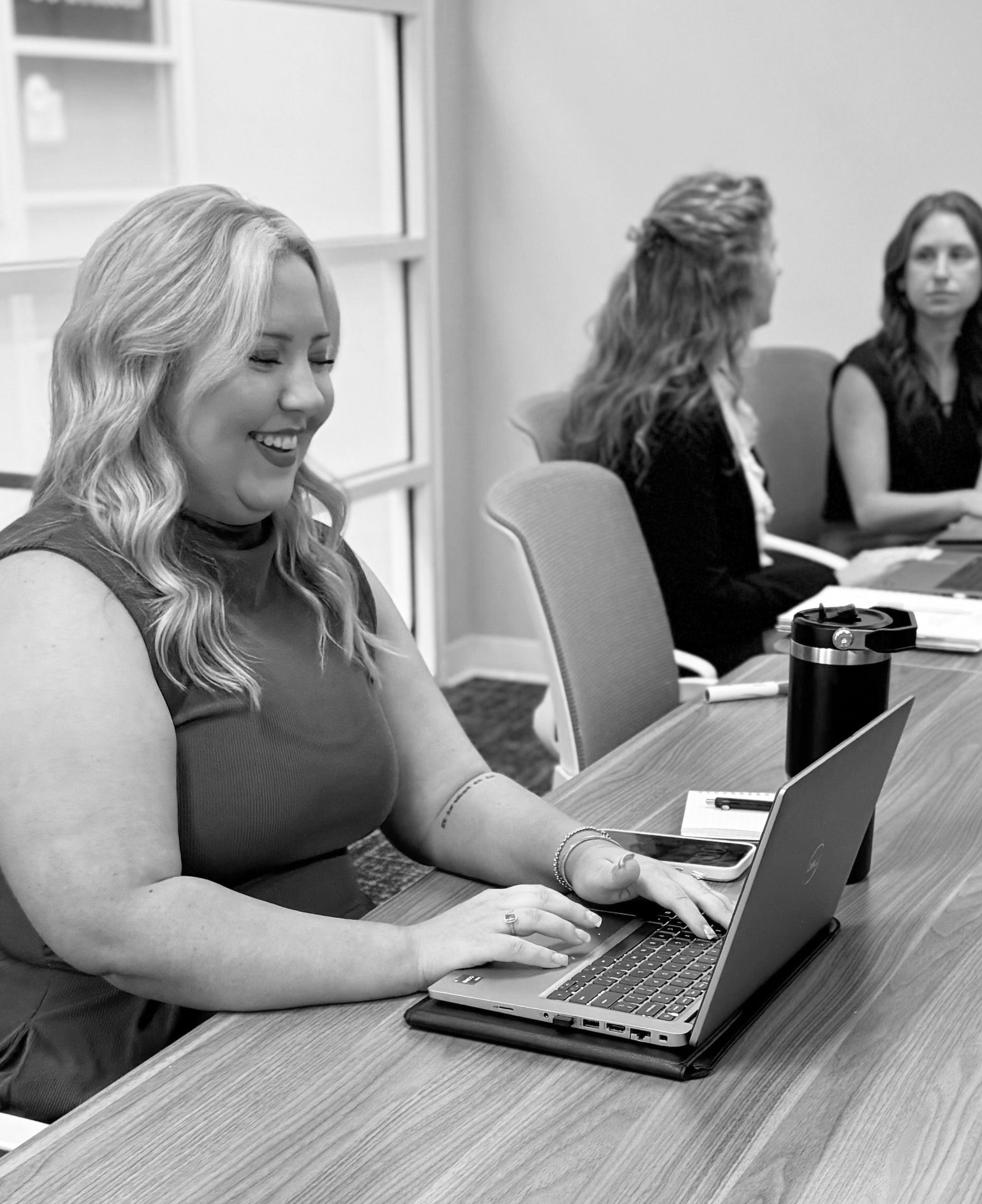 Woman typing on a laptop at a conference table, with two colleagues seated in the background.