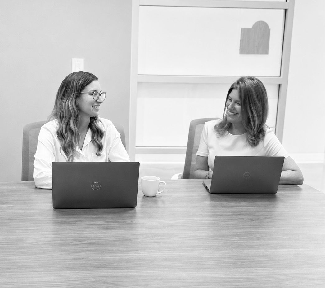 Two people seated at a conference table, smiling and talking with laptops open in a meeting room.
