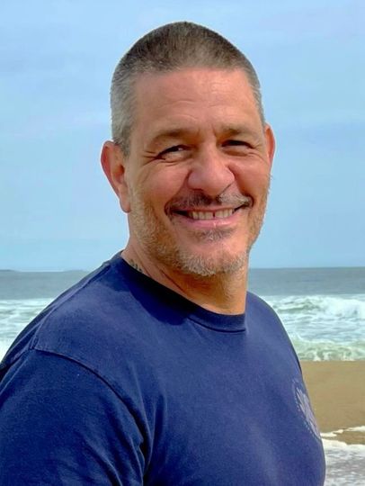 Man with short hair and stubble smiles at the camera on a beach with ocean waves.