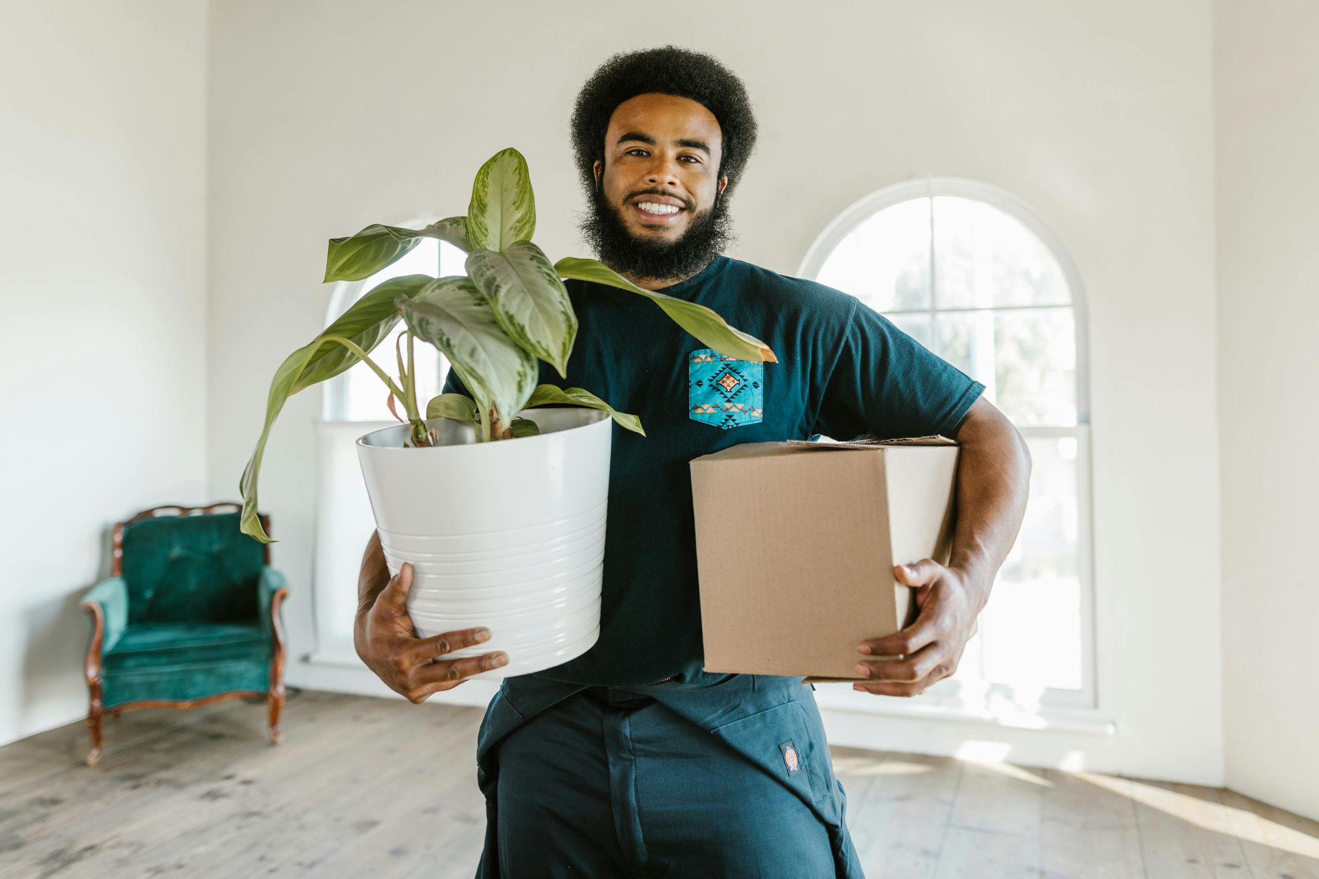 Man with beard smiles while holding a potted plant and a cardboard box in an empty room with a window.