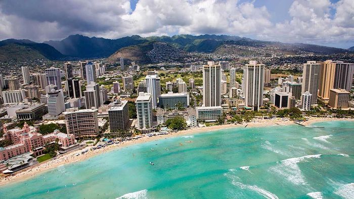 Aerial view of a coastal city with high-rise buildings, turquoise bay, sandy beach, and mountains in the background