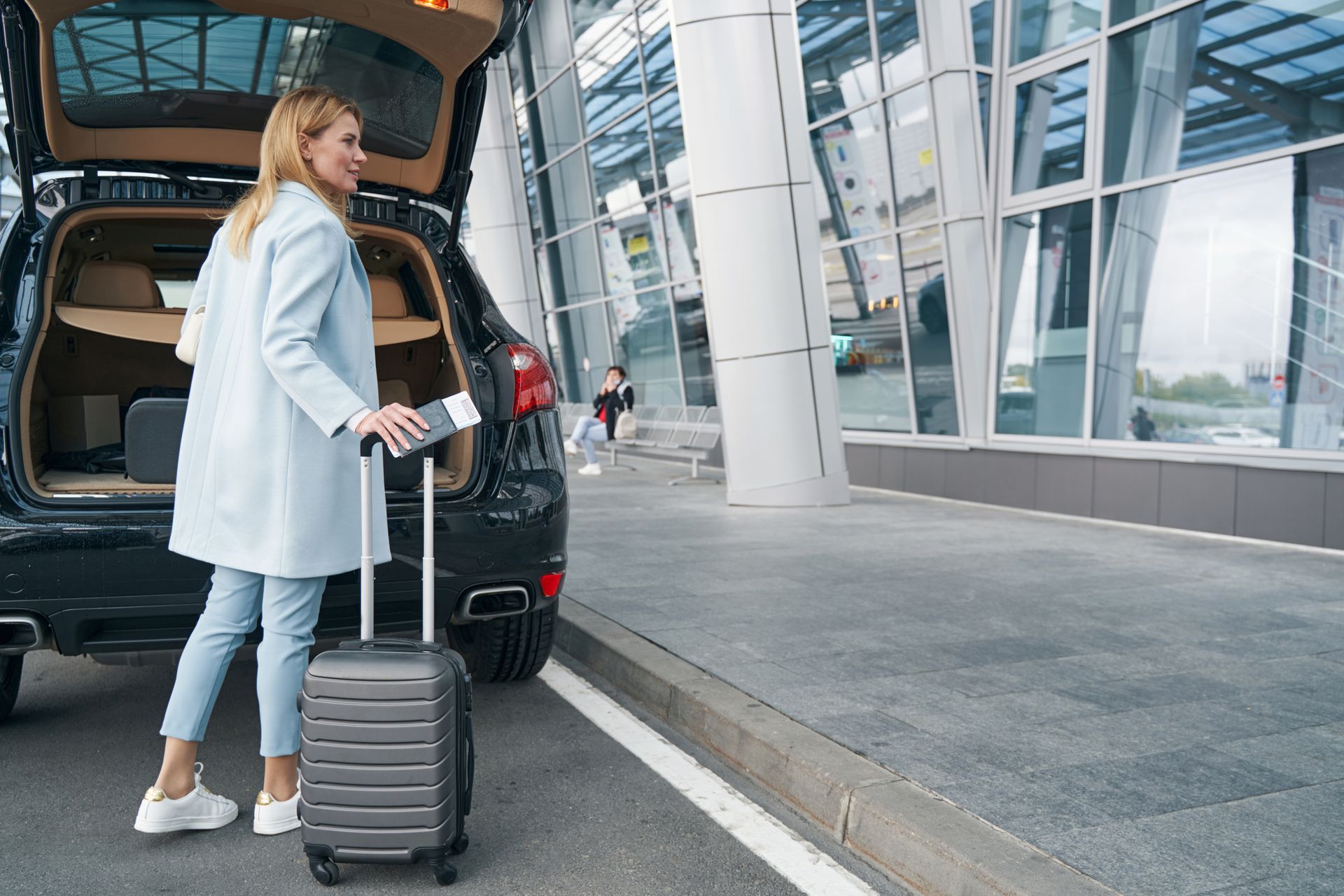 A woman with a suitcase is standing next to a car with the trunk open.