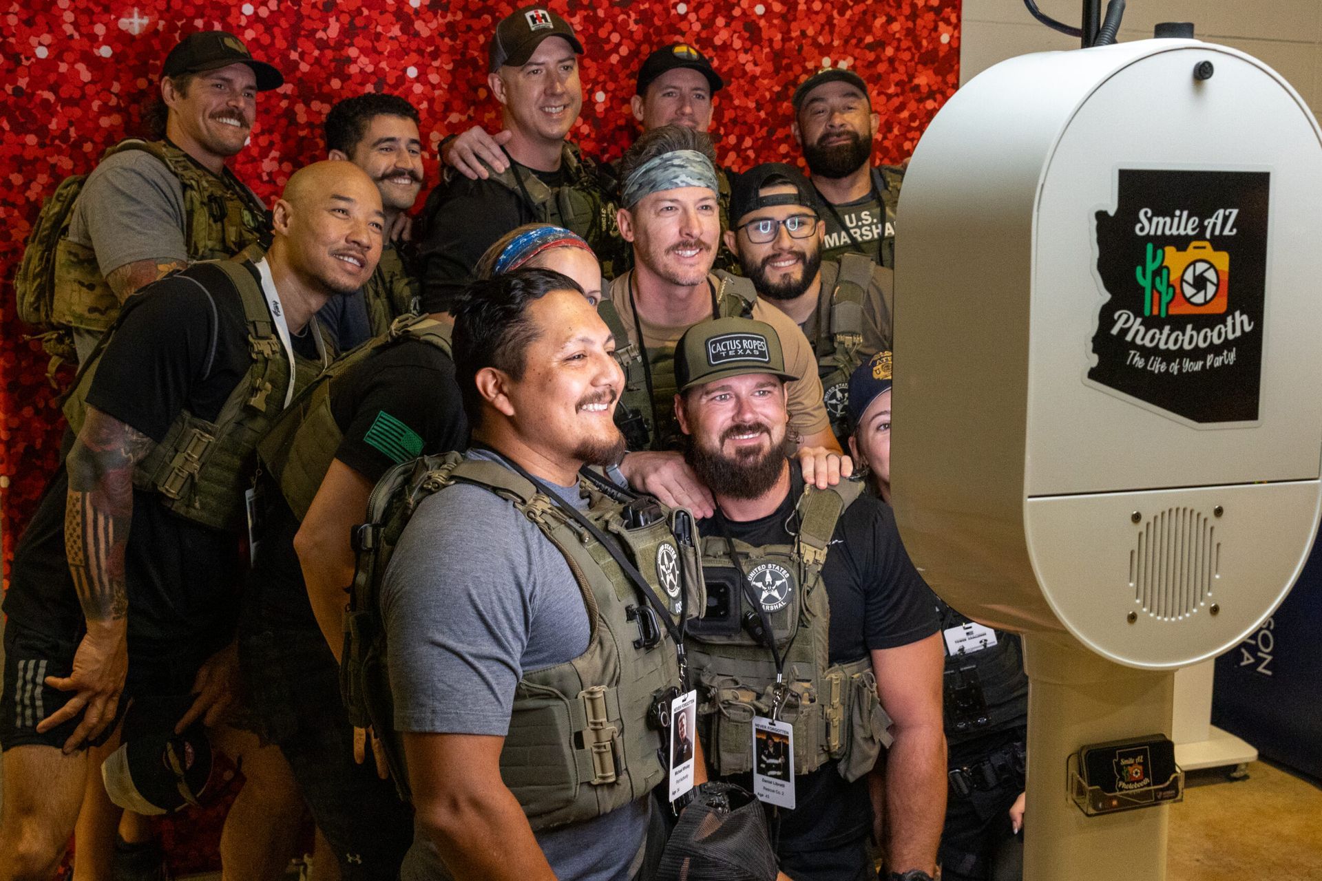 Group of men in tactical vests smiling for a photo at a photobooth with a red glitter backdrop.