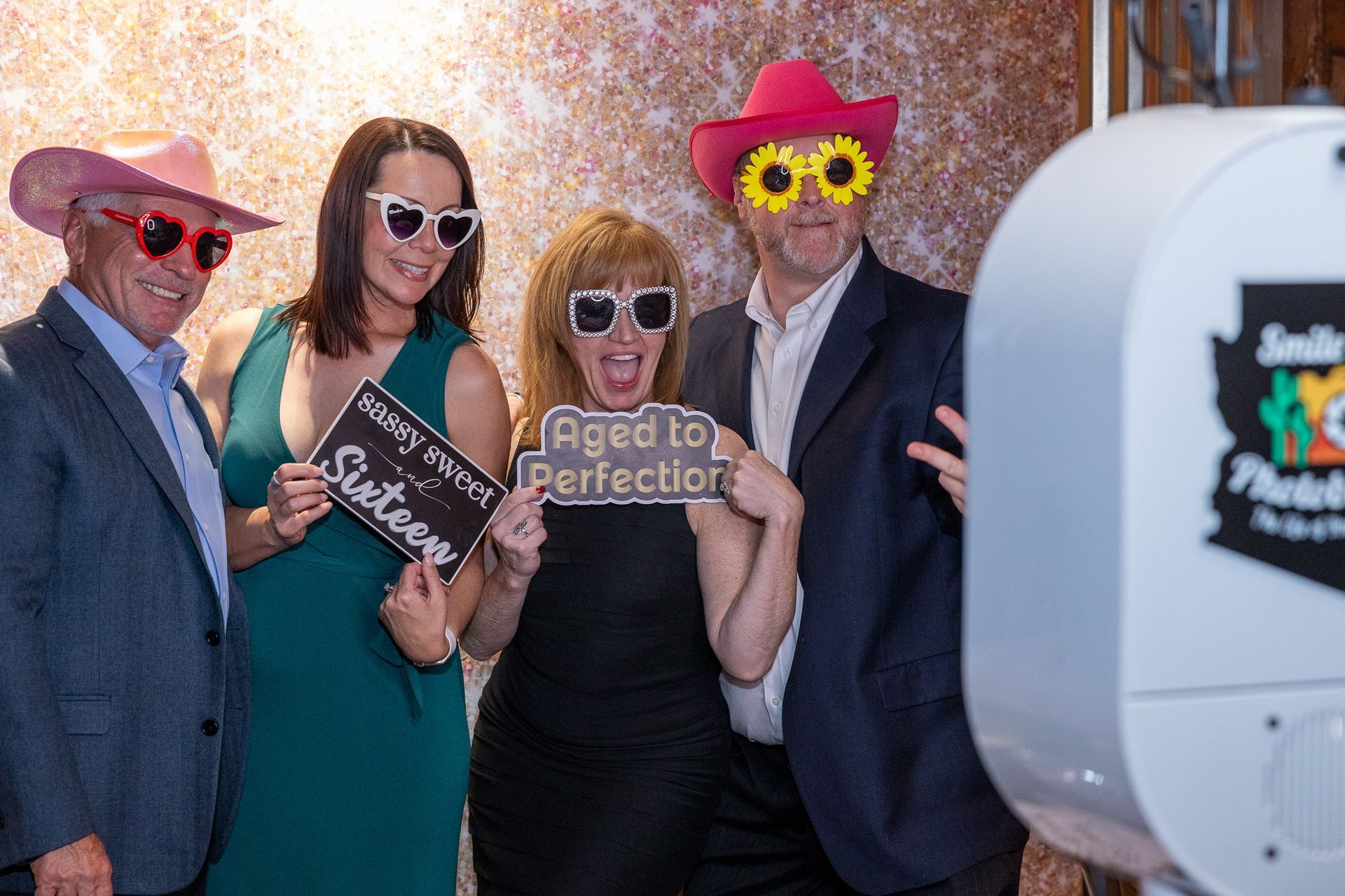 Four people pose in a photo booth, wearing sunglasses and hats. One holds a sign that says