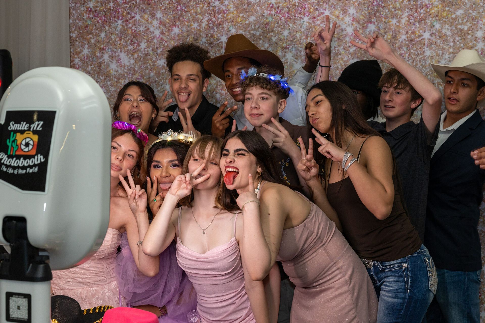 Group of people posing for photo booth. Some making peace signs, others smiling, in front of a colorful backdrop.