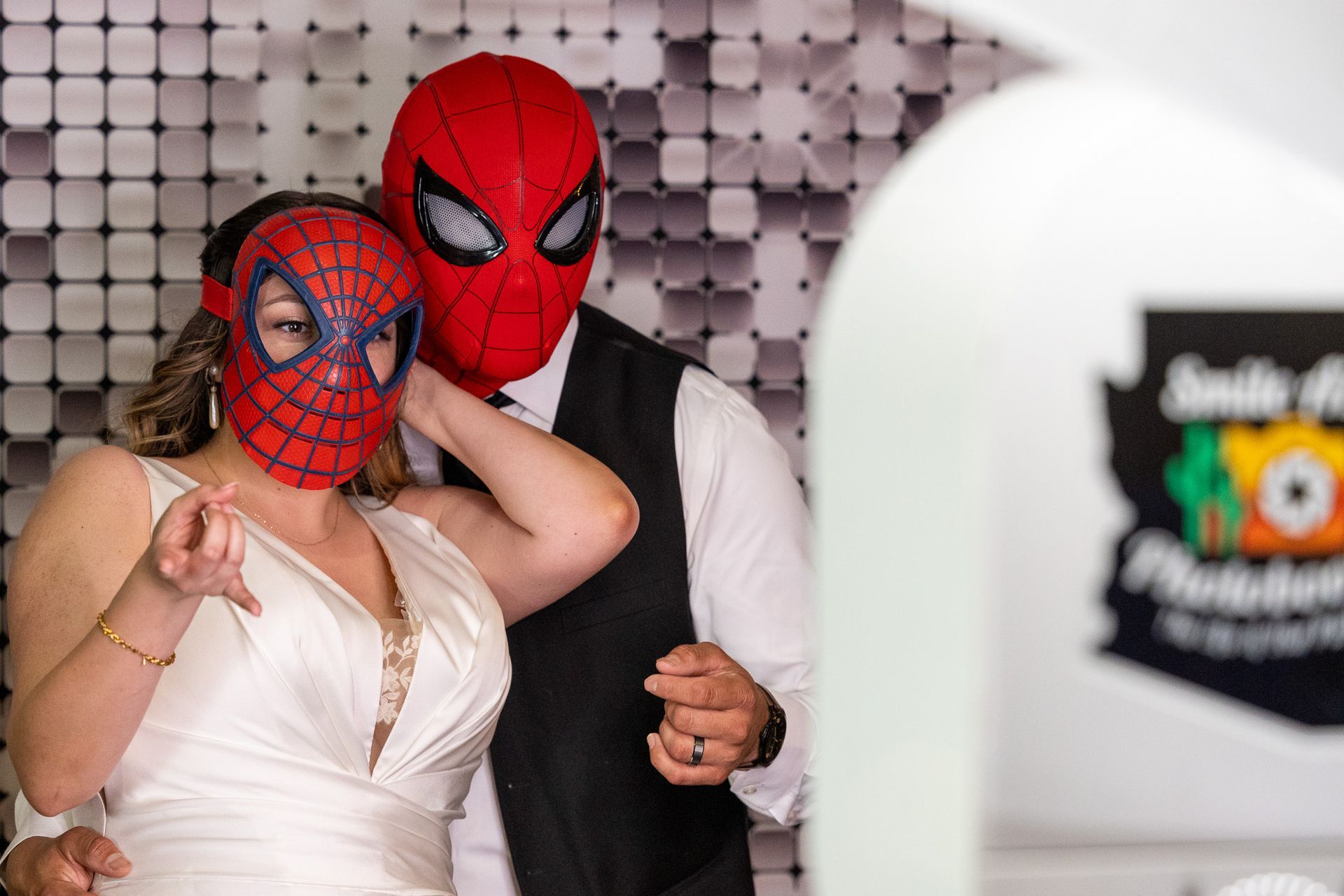Couple wearing Spider-Man masks in photo booth; bride points, groom stands behind.