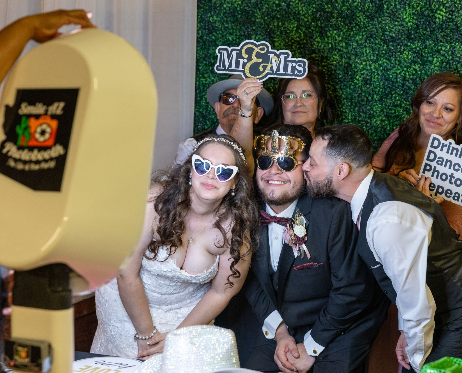 Wedding photo booth with bride, groom, and guests smiling, holding props. Green backdrop, warm lighting.