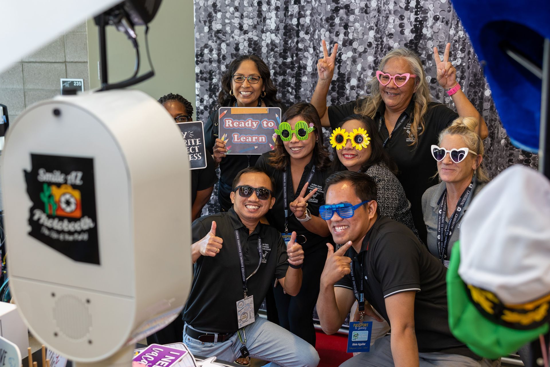 People posing for photo in front of a backdrop, some wearing novelty glasses and holding signs, by a photobooth.