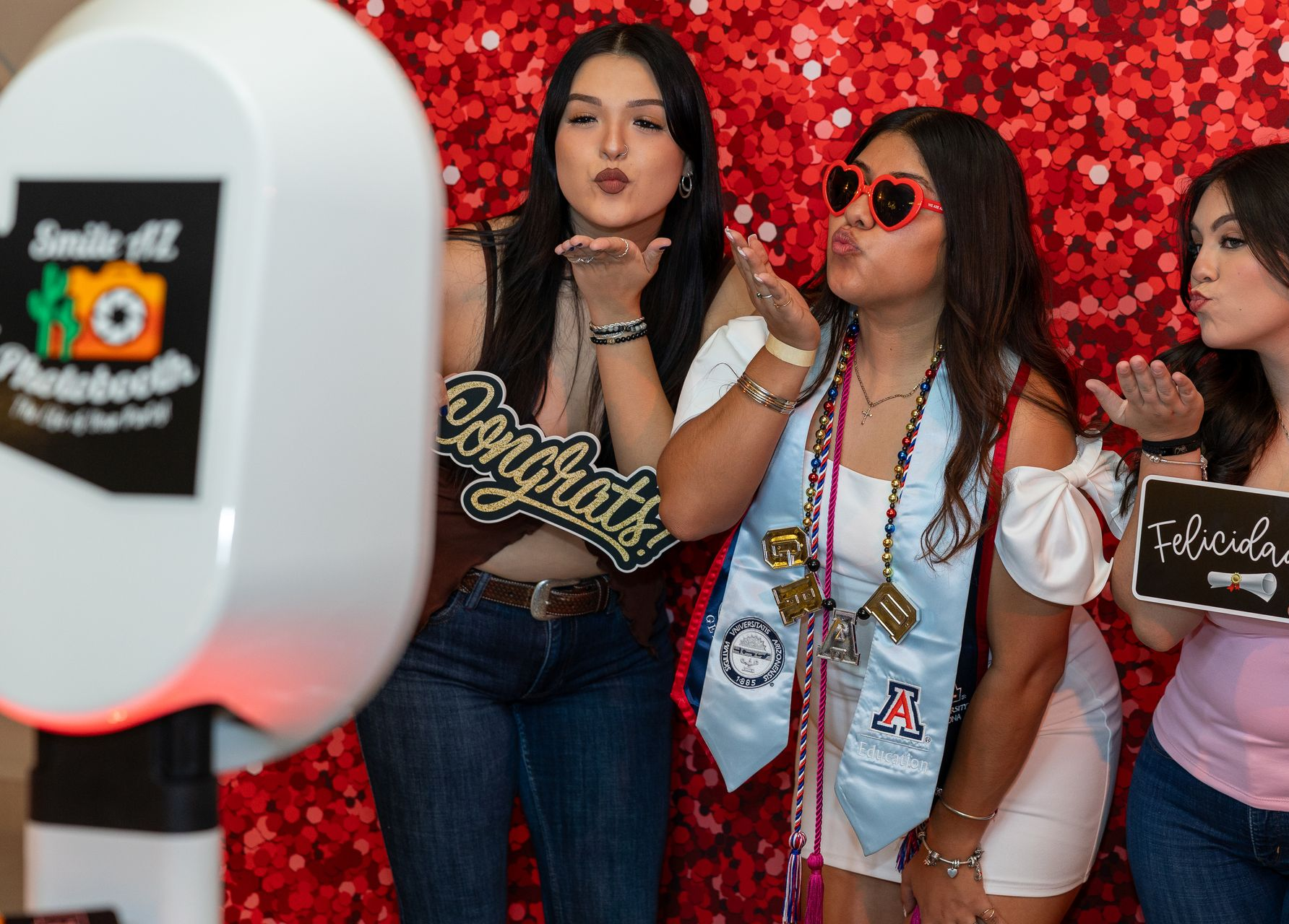 Three women pose in front of a photo booth, blowing kisses; red glitter backdrop.