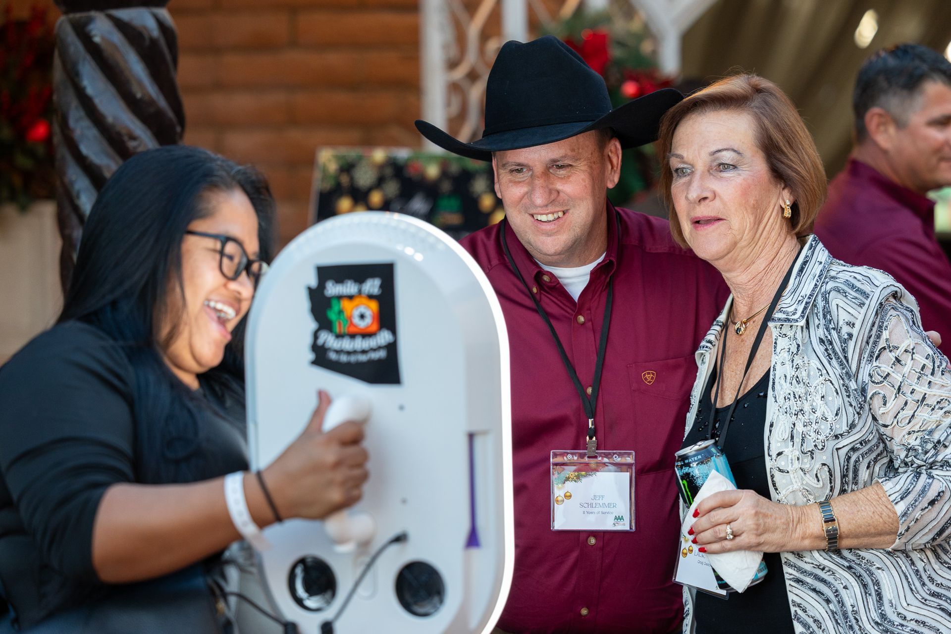 People smiling at a photo booth; one in cowboy hat, another with a drink, and one operating it.