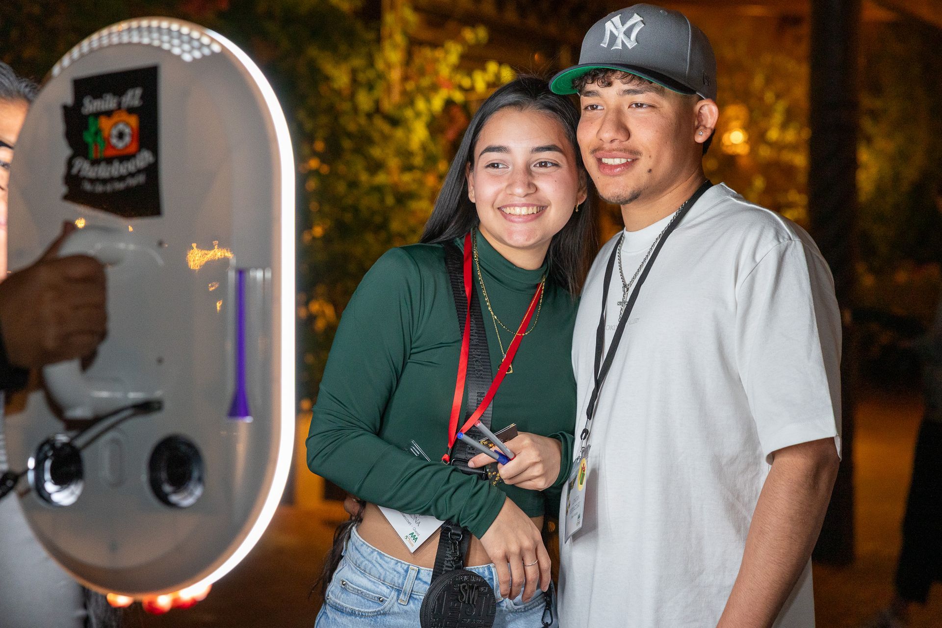 Two people smile posing beside a photo booth at an outdoor event at night.