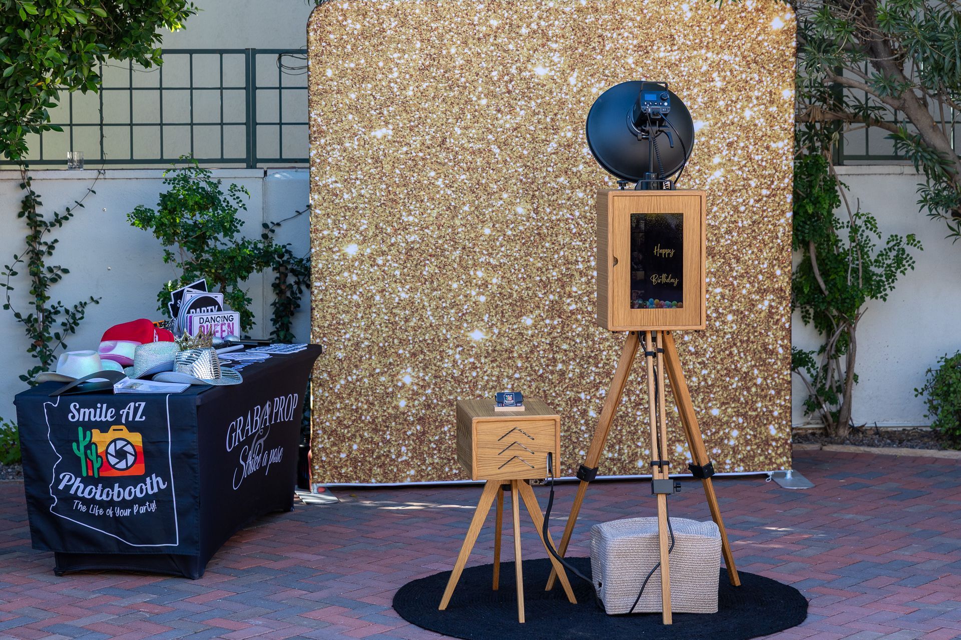 Photo booth with gold glitter backdrop and prop table. Outdoor setting.