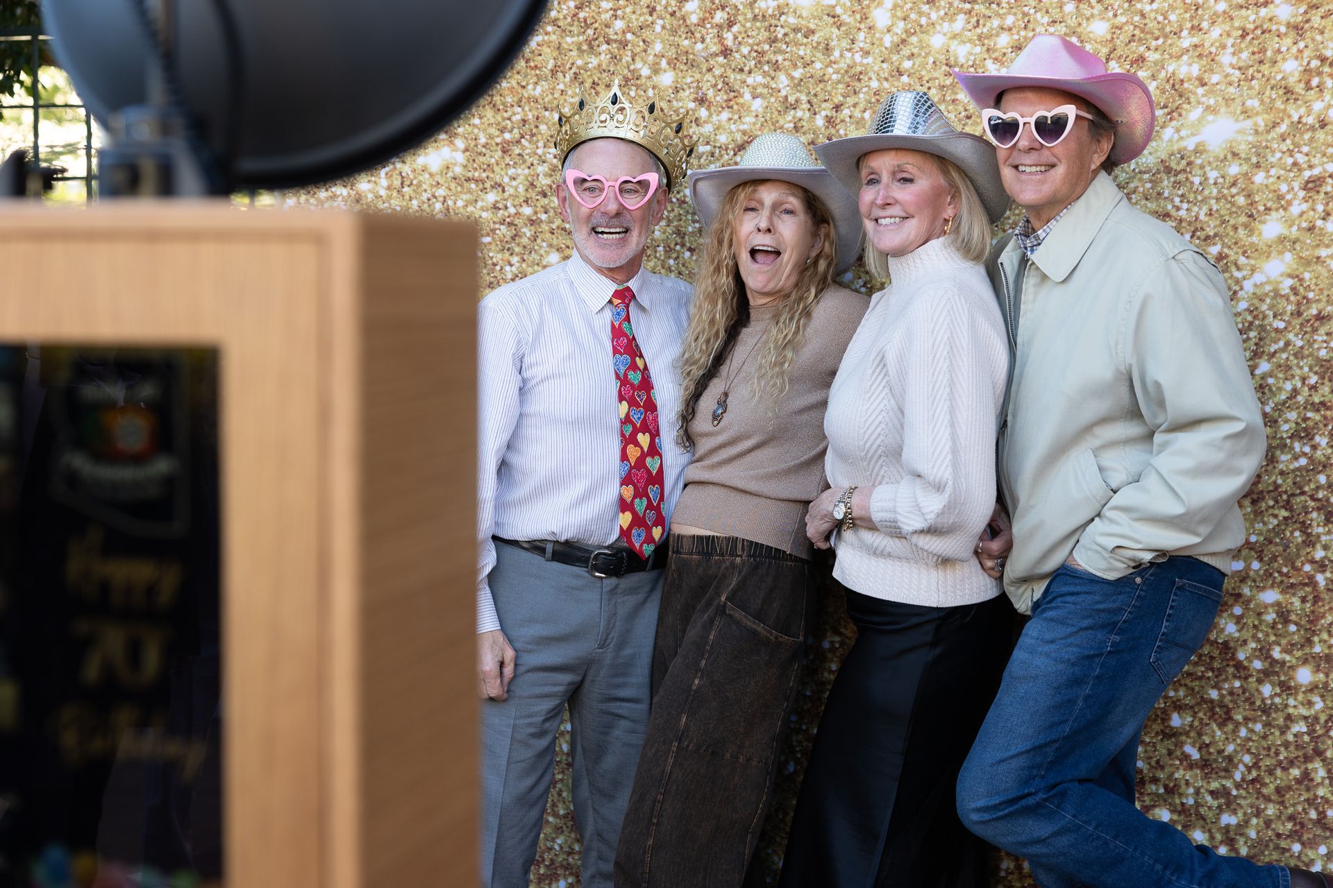 Four people pose with props in front of a gold glitter backdrop at a photo booth.