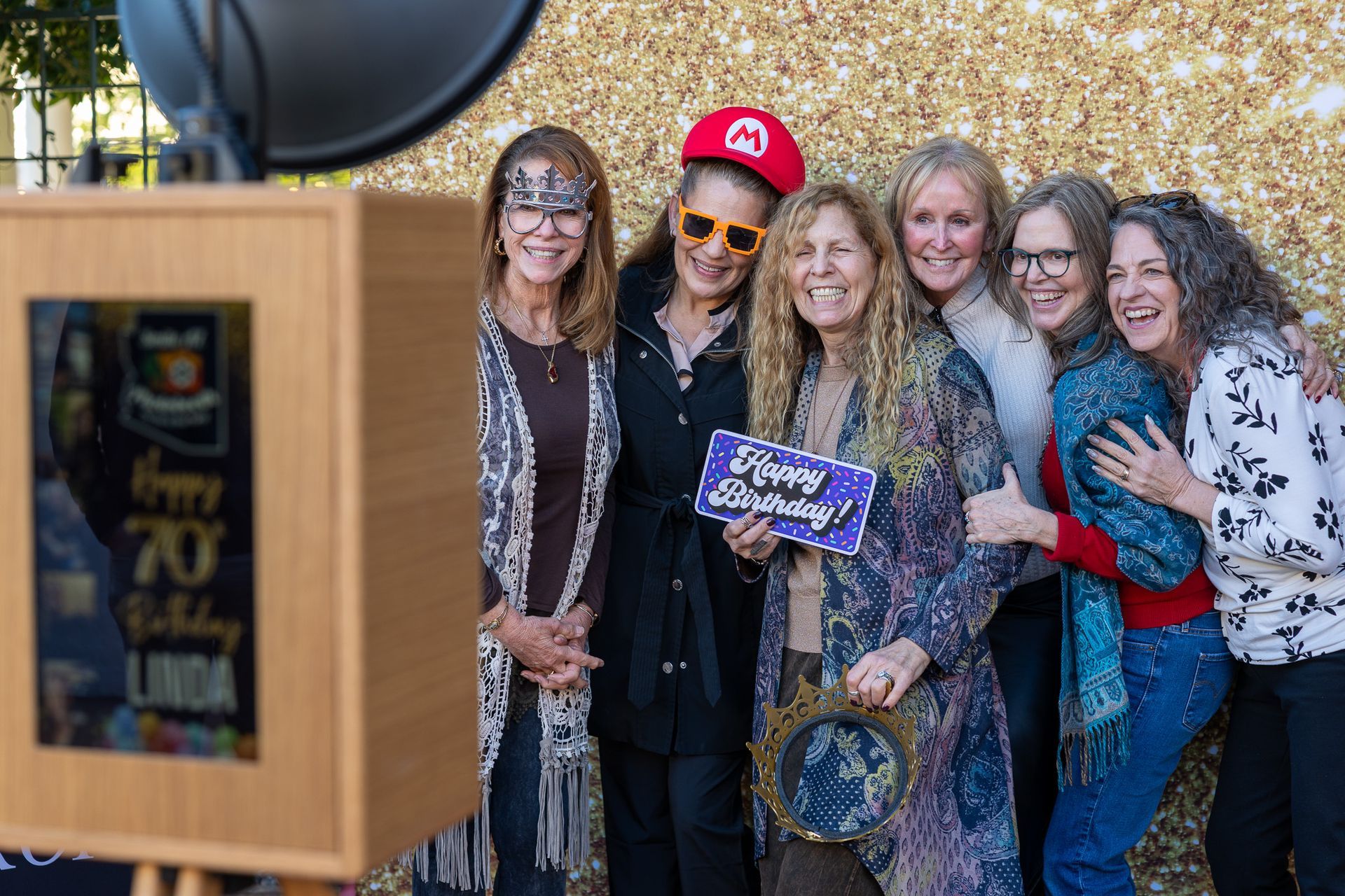 Group of women pose at a photo booth with prop celebrating a 70th birthday.