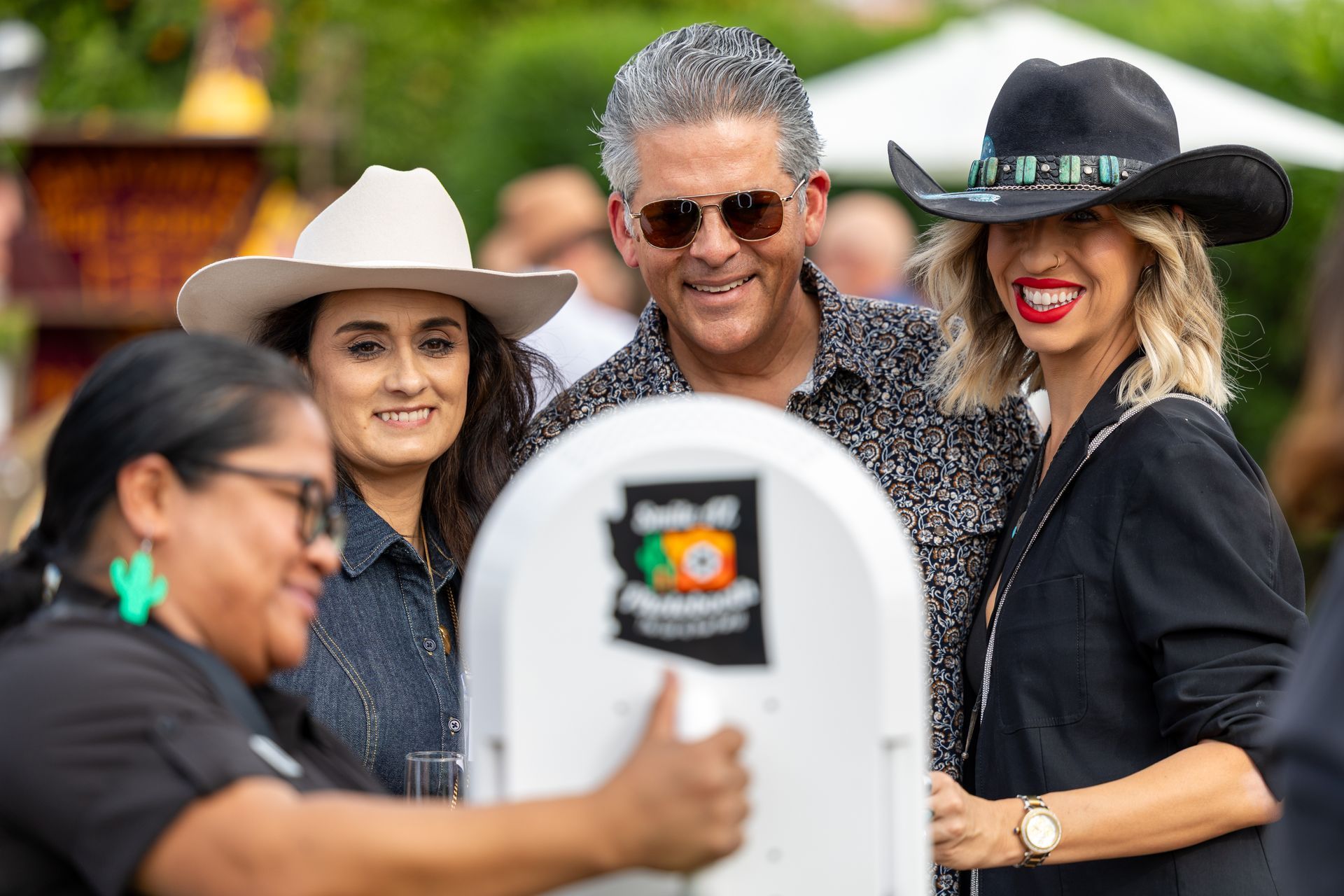 People at a photo booth; a woman operates the machine while a man and another woman look on. Outdoors.