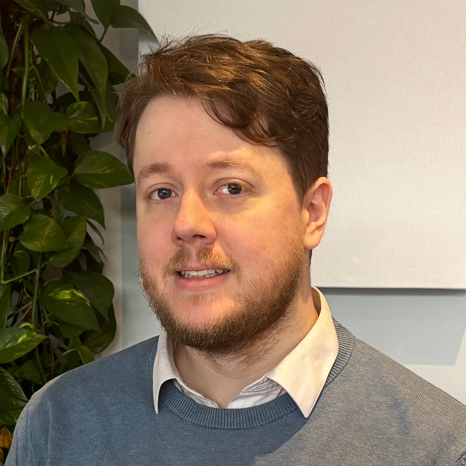 Man with light skin and a short brown beard, wearing a blue sweater and white collared shirt, smiles near green plants.