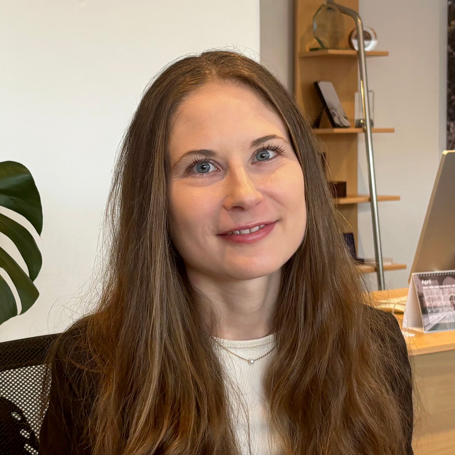 Woman with long brown hair smiling, indoors. Wearing a white top, black blazer, near a desk.