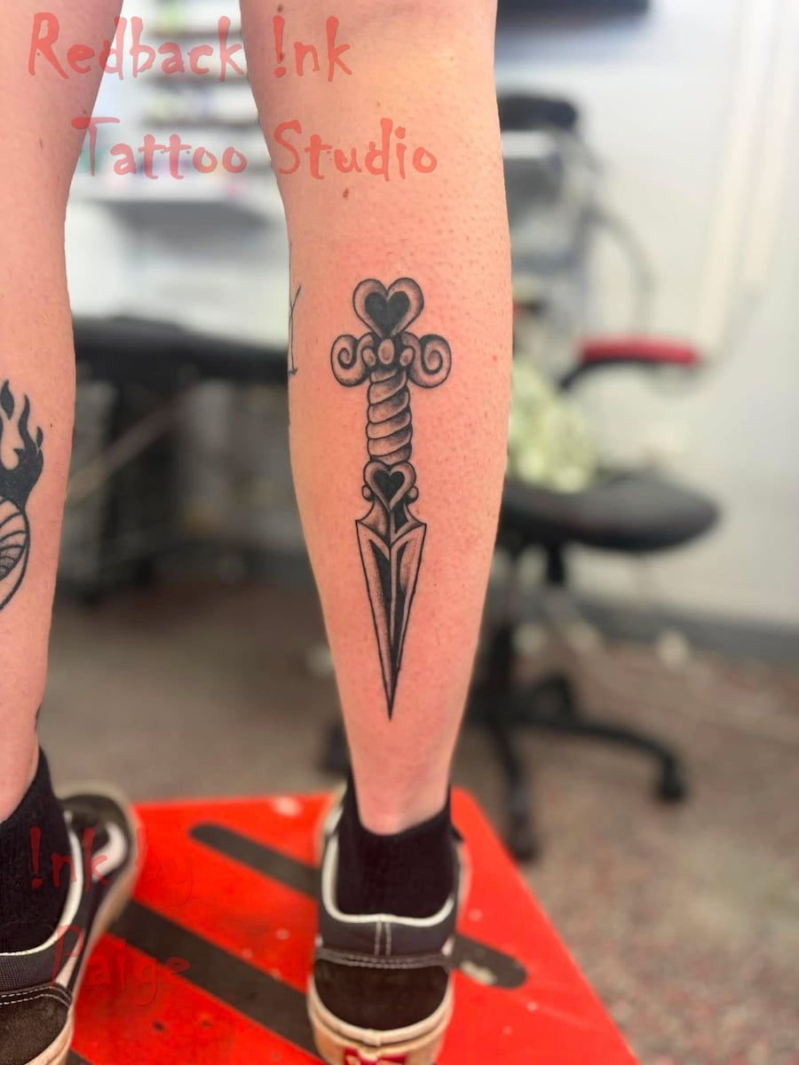 Tattooed Lower Leg With Black Dagger Design and Red Watermark Text — Redback !nk Tattoo Studio in Kawana, QLD