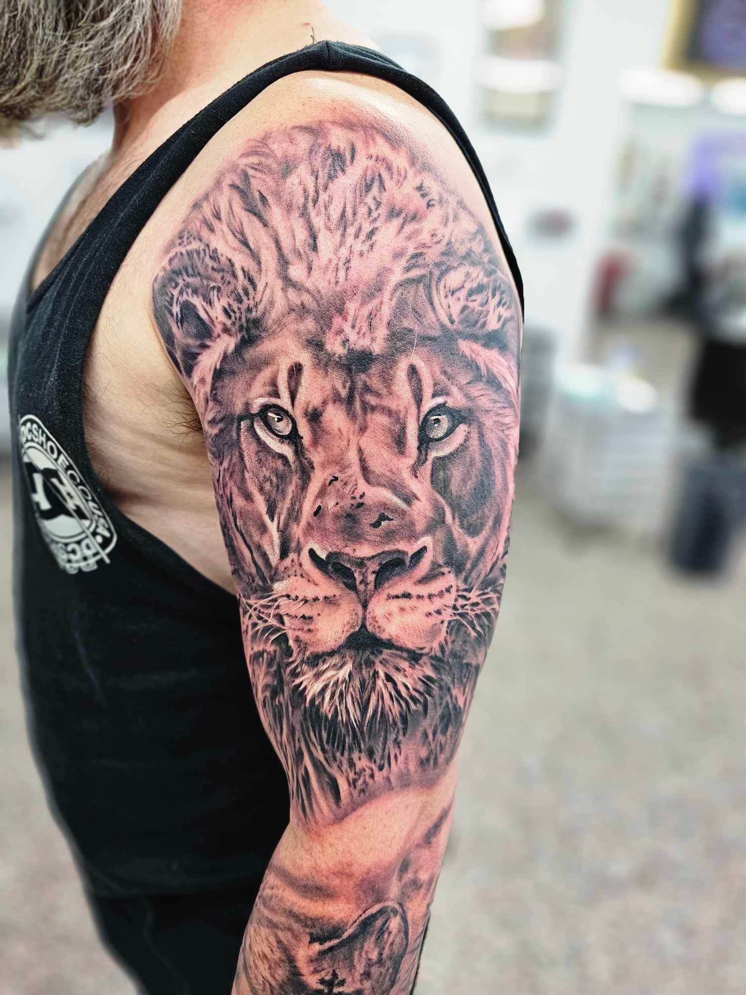 Black and White Lion Tattoo on a Man's Upper Arm; He's Wearing a Black Tank Top — Redback !nk Tattoo Studio in Kawana, QLD
