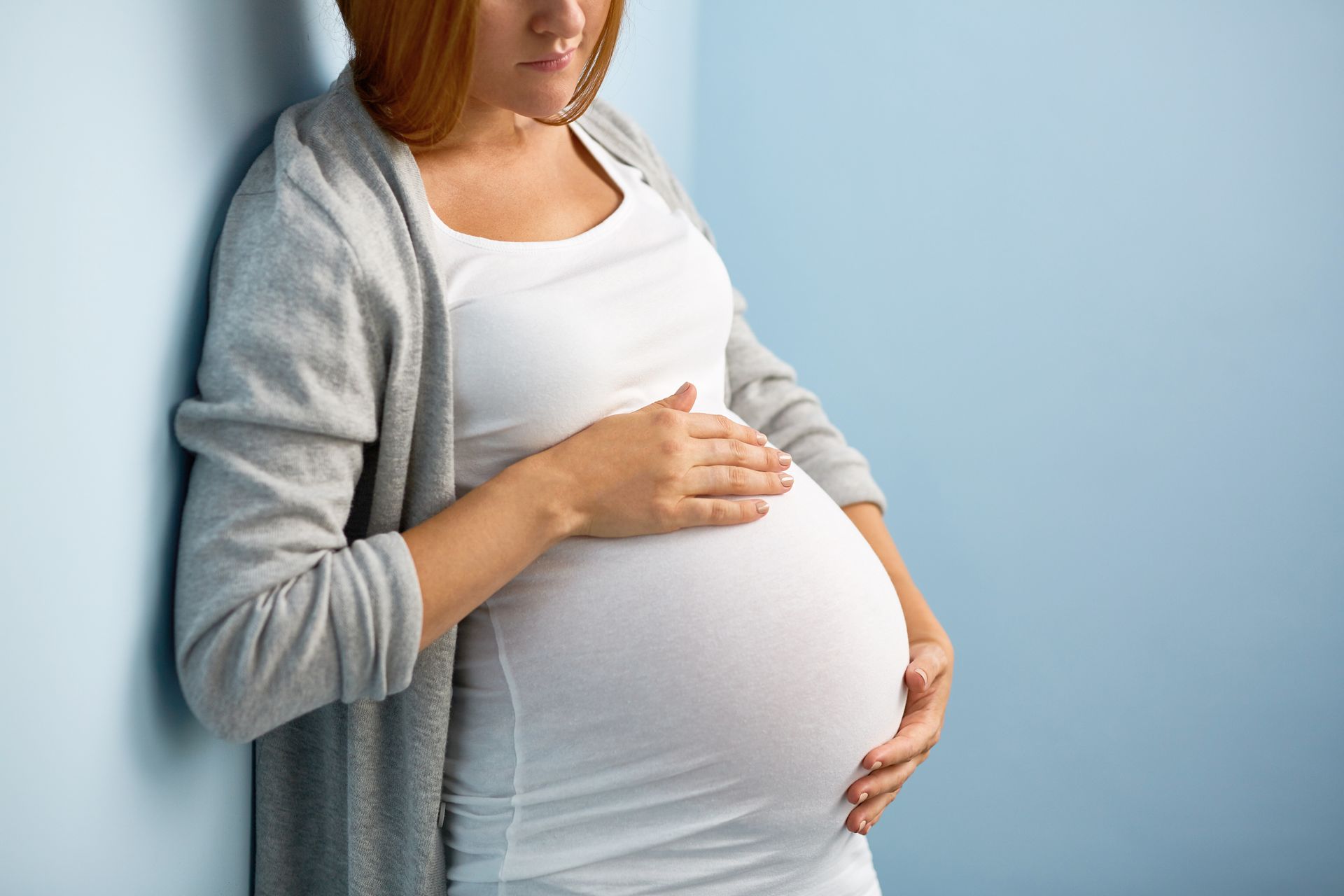 Pregnant woman in a white dress and gray cardigan, holding her belly, leaning against a blue wall.