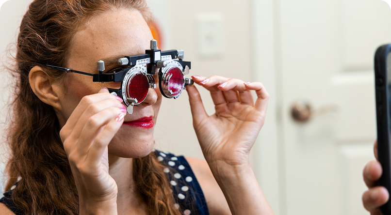 Woman in an eye exam, looking through phoropter with red lenses.
