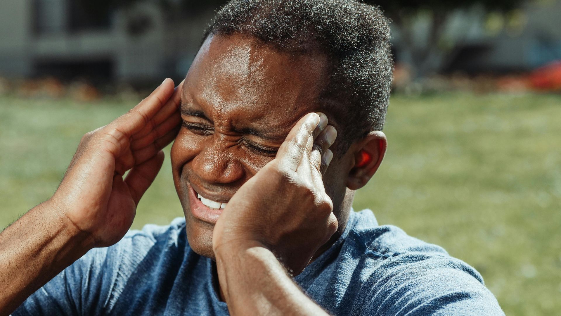 Man holding his temples with eyes shut, grimacing outdoors on a sunny day