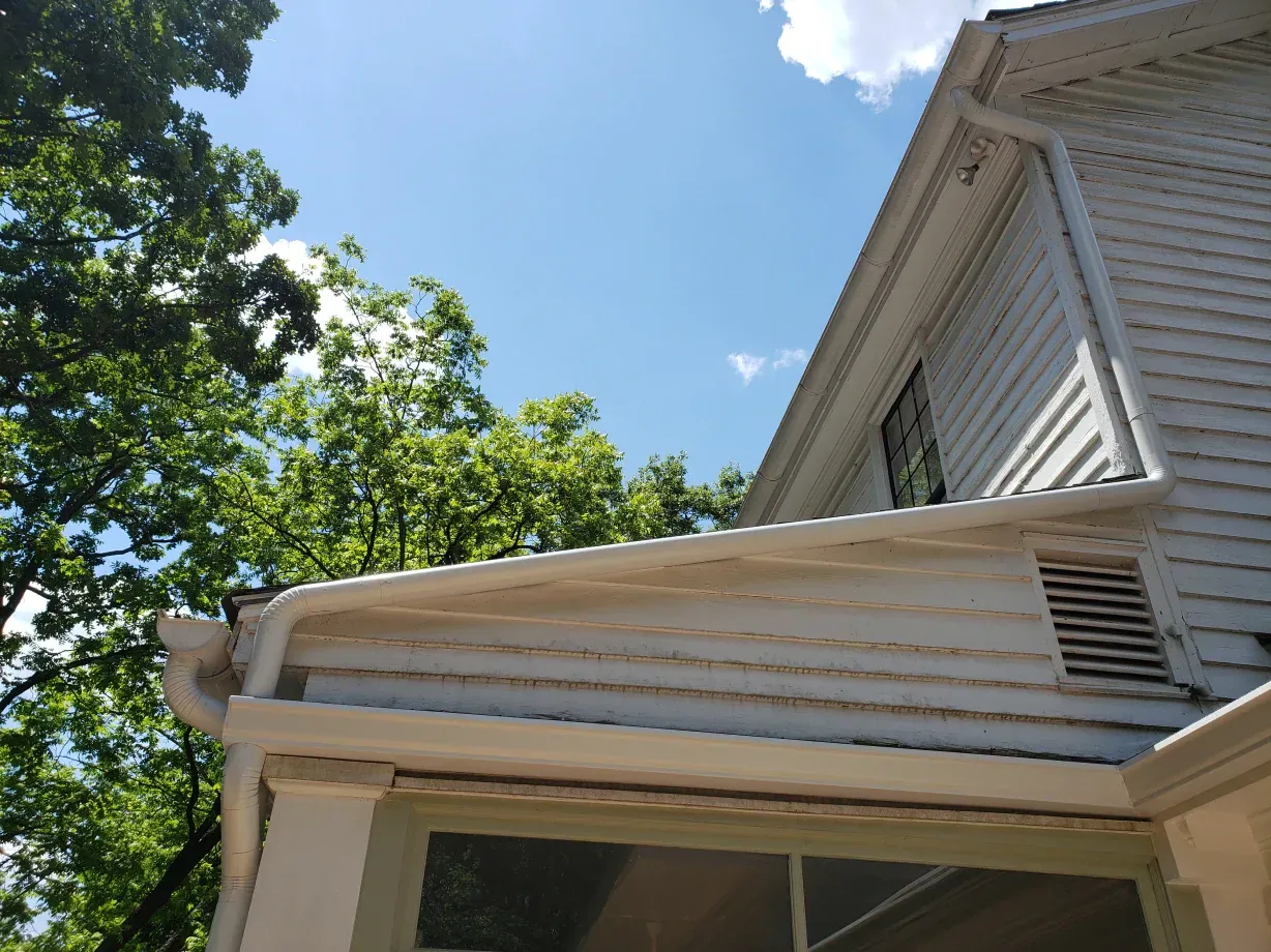 White house siding with a gutter and vent, under a blue sky with trees.