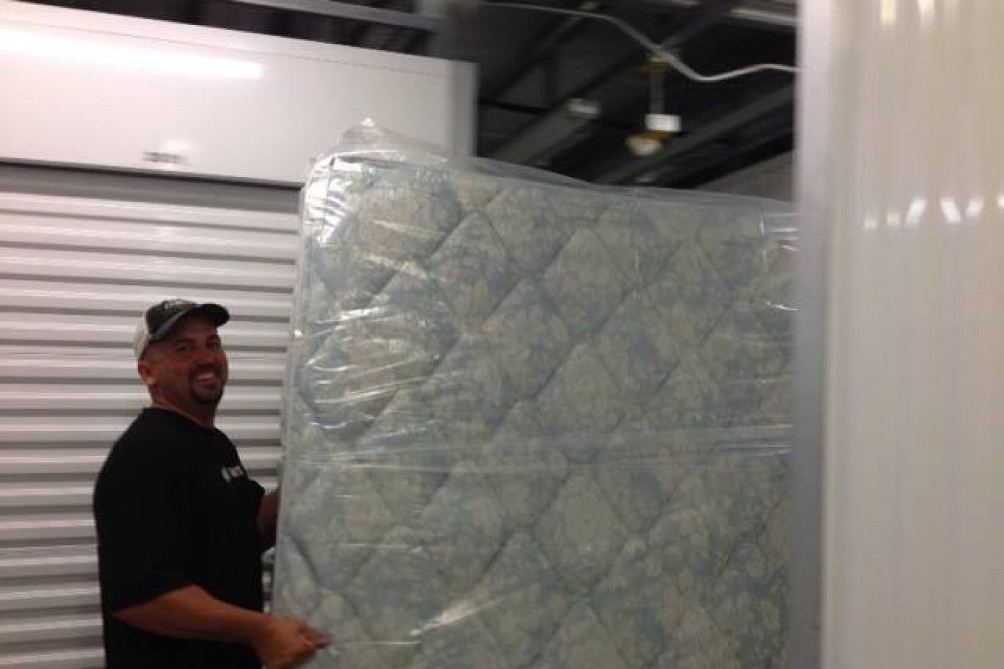 Man holding a plastic-wrapped mattress in a storage unit, smiling.