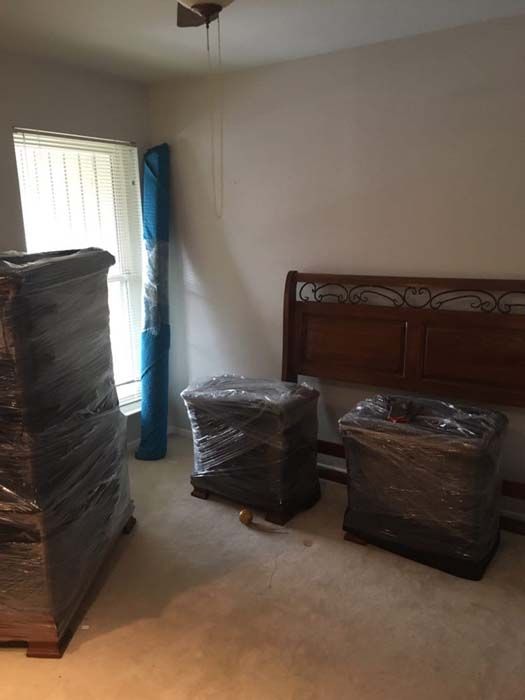 Bedroom with furniture wrapped in plastic, including a bed frame, nightstands, and a tall dresser. A rolled-up rug stands upright.