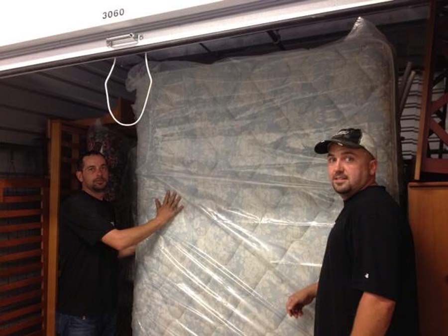 Two men in a storage unit holding a large, wrapped mattress.