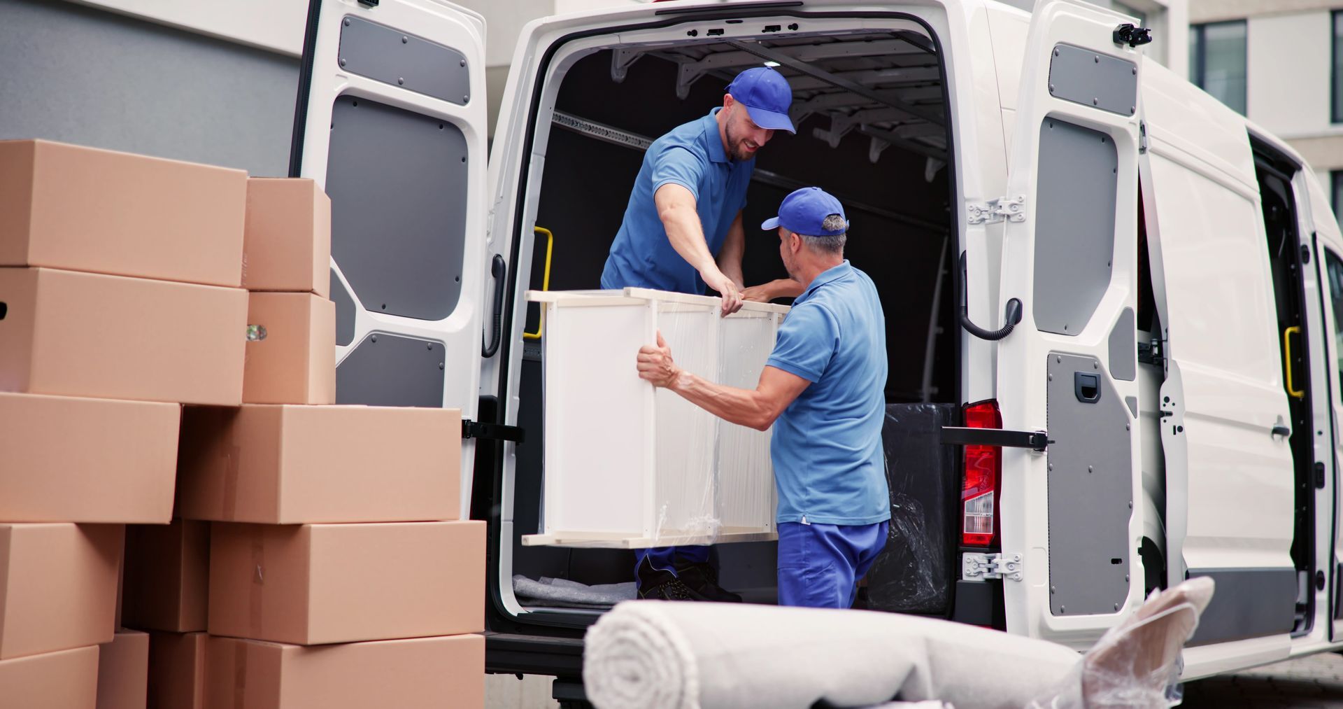 Two movers loading a white container into a van, surrounded by cardboard boxes.