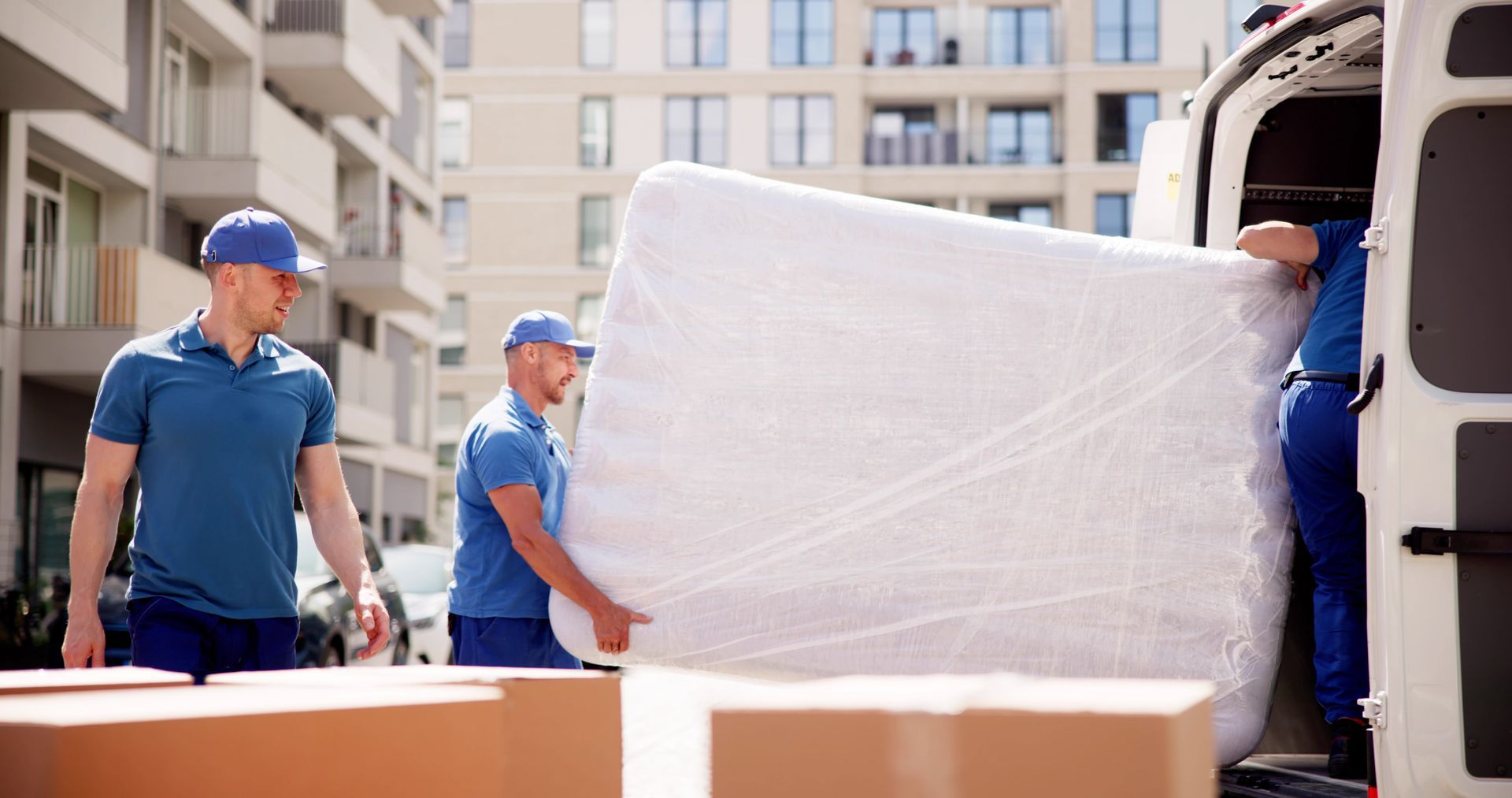 Movers in blue shirts and caps carrying a wrapped mattress to a van near an apartment building.