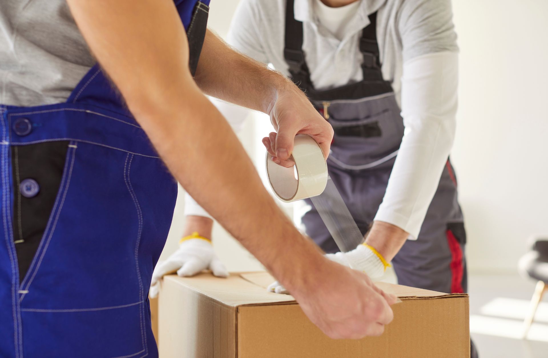 Two movers sealing a cardboard box with tape, wearing work overalls and gloves.