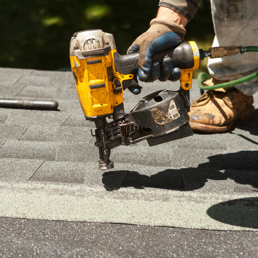 Worker using a yellow roofing nail gun on gray shingles beside a rooftop safety harness