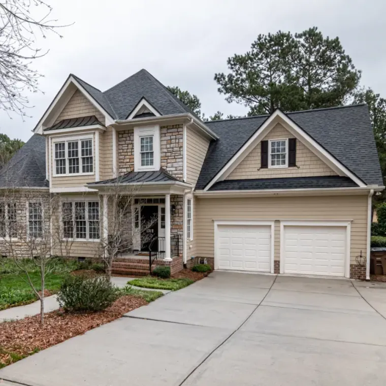 Suburban two-story house with beige siding, gray roof, and a double garage beside a curved driveway.
