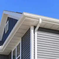 Close-up of a gray house corner with white soffit, gutter, and blue siding against a clear blue sky