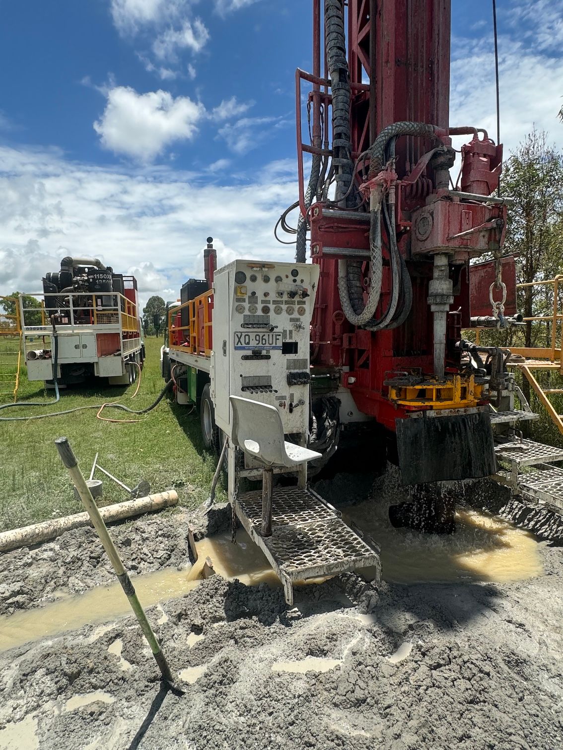 Drilling rig in operation, red and white machinery, on a muddy ground with two service trucks nearby — Kowaltzke Drilling Services in Gatton, QLD