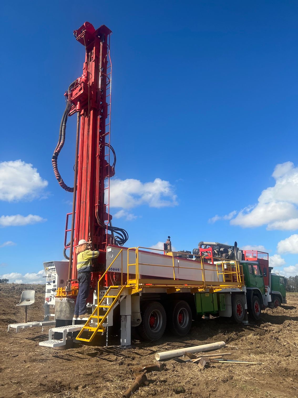 Red drilling rig on wheels, in a field. A worker is present. Blue sky — Kowaltzke Drilling Services in Dalby, QLD