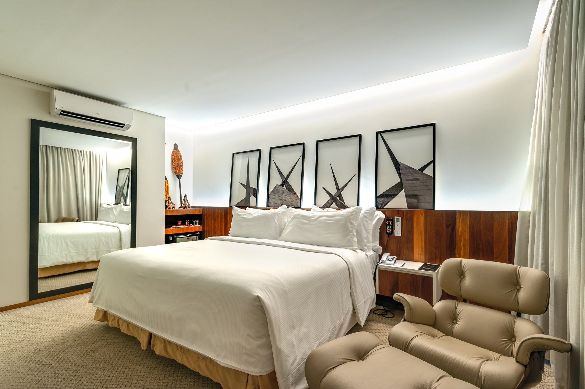 Quarto de hotel moderno com cama king-size, guarda-roupa espelhado, obras de arte na parede e uma poltrona bege.
