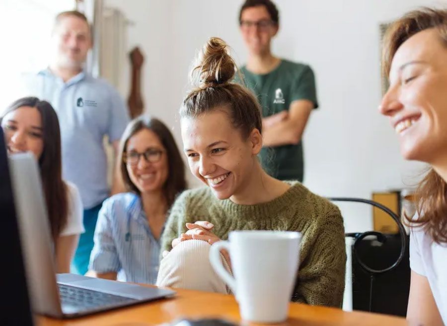 Group of smiling people gather around a laptop, likely collaborating in a bright room.