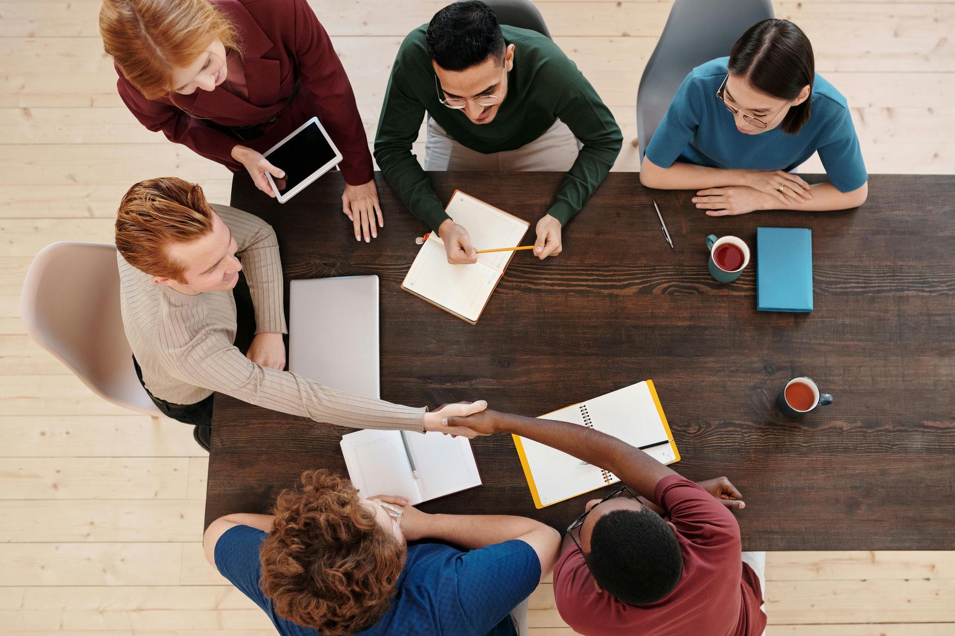People gathered around a wooden table in a meeting, writing on papers with laptops, notebooks, and coffee cups.