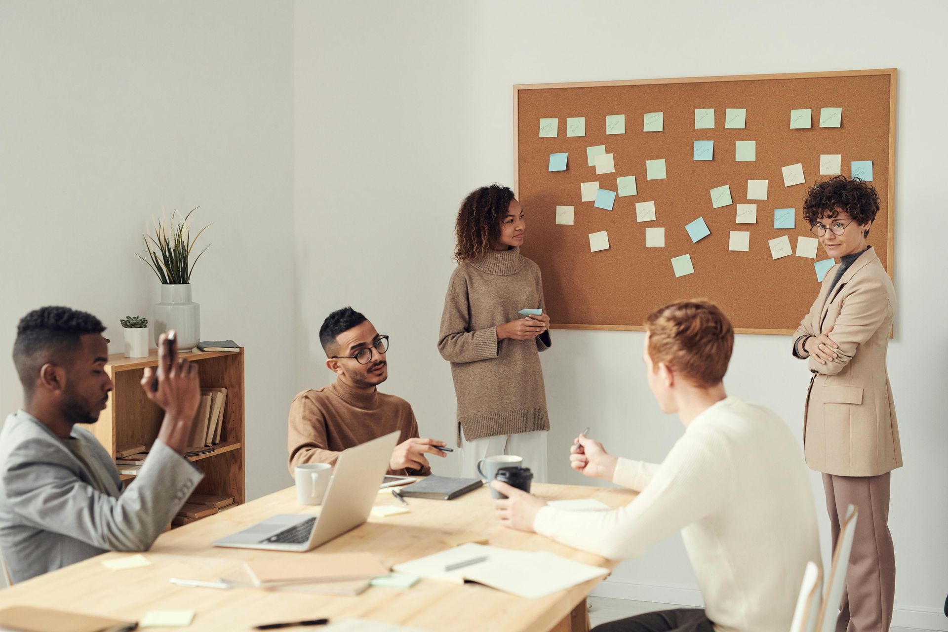 Business team in a meeting. A man in a suit stands in front of a glass wall with sticky notes, pointing while others look on.