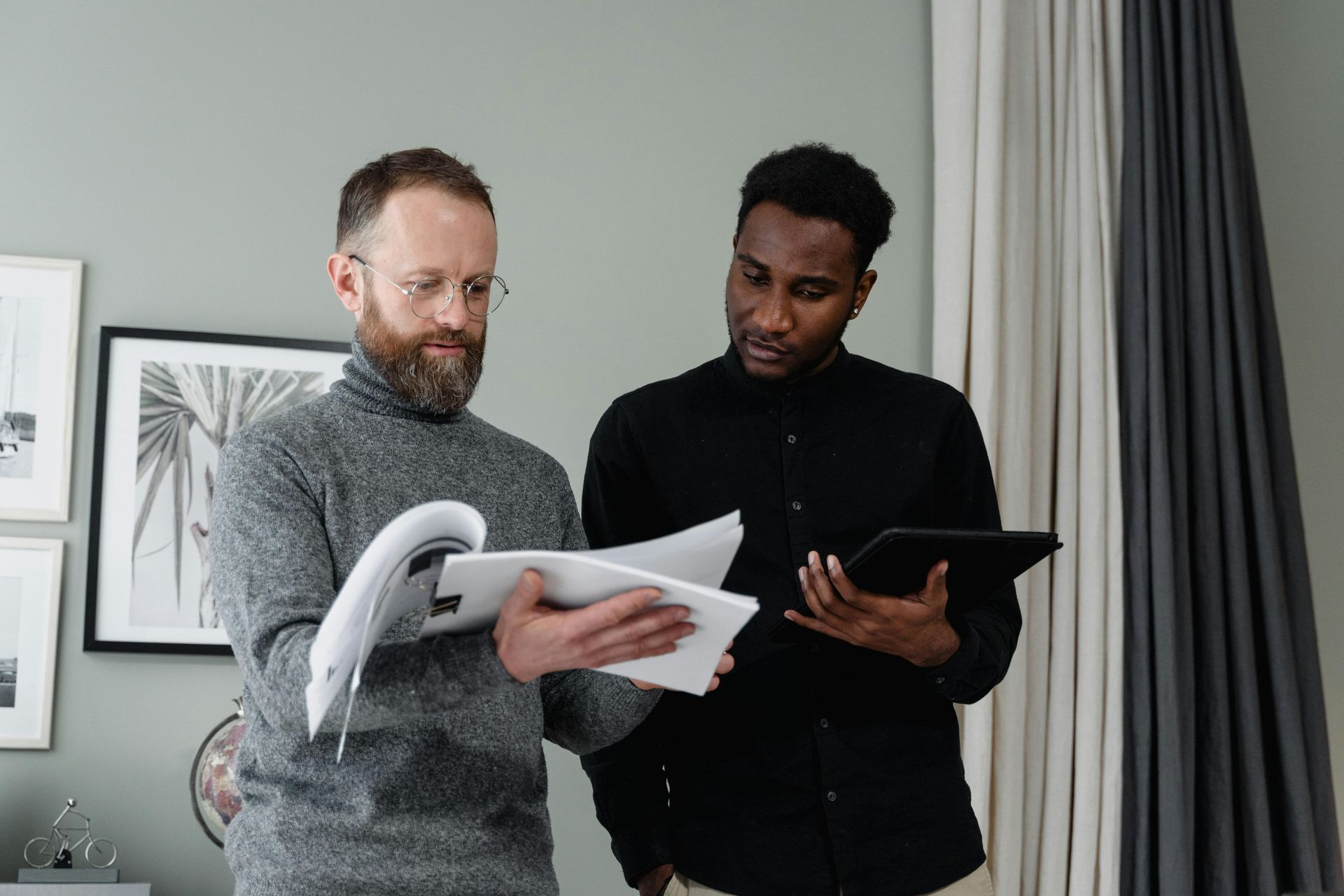 Two men, one with a beard holding papers, the other with a tablet, discussing documents.