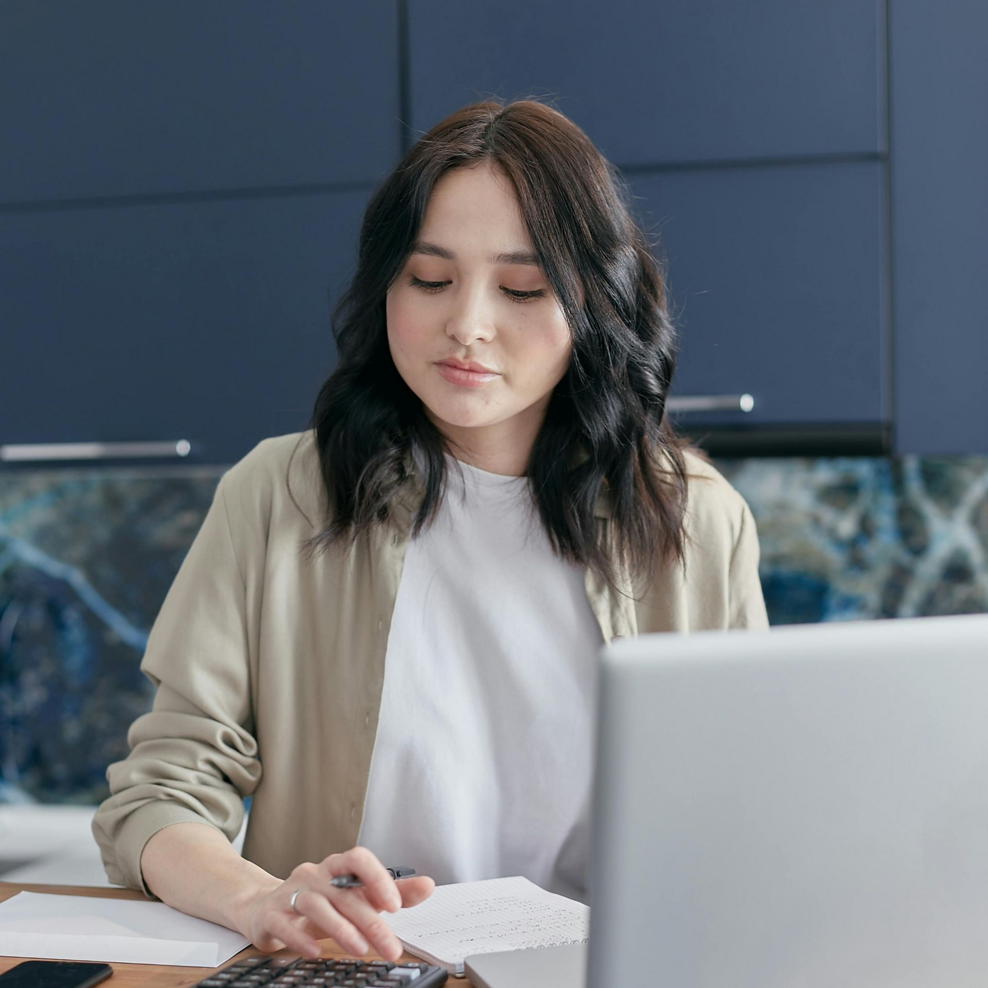 Woman with dark hair working on finances, using calculator and laptop in kitchen.