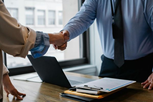 Two people in business attire shaking hands over a desk with a laptop and documents. Indoor setting with natural light.