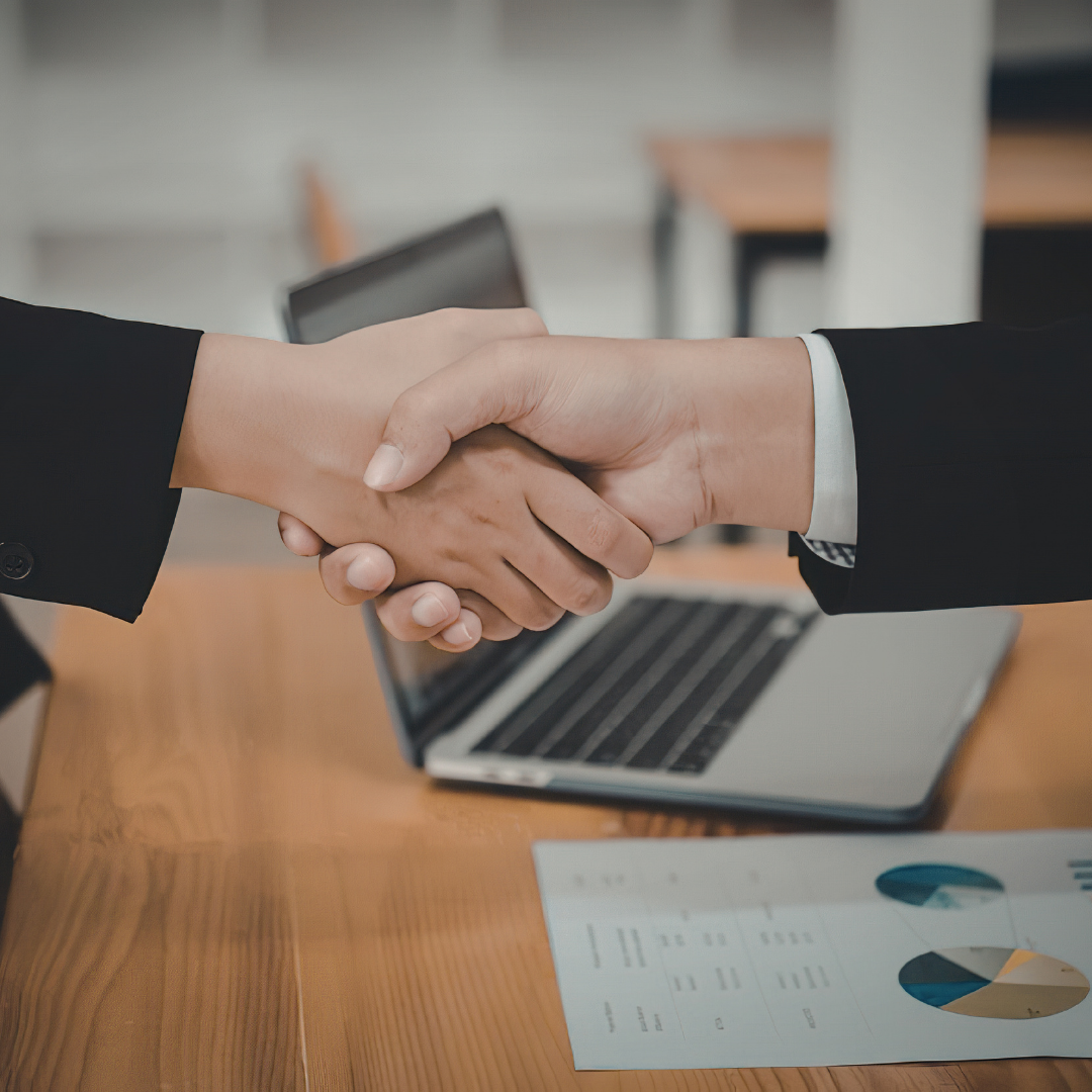 Two people shaking hands over a laptop and business chart on a desk