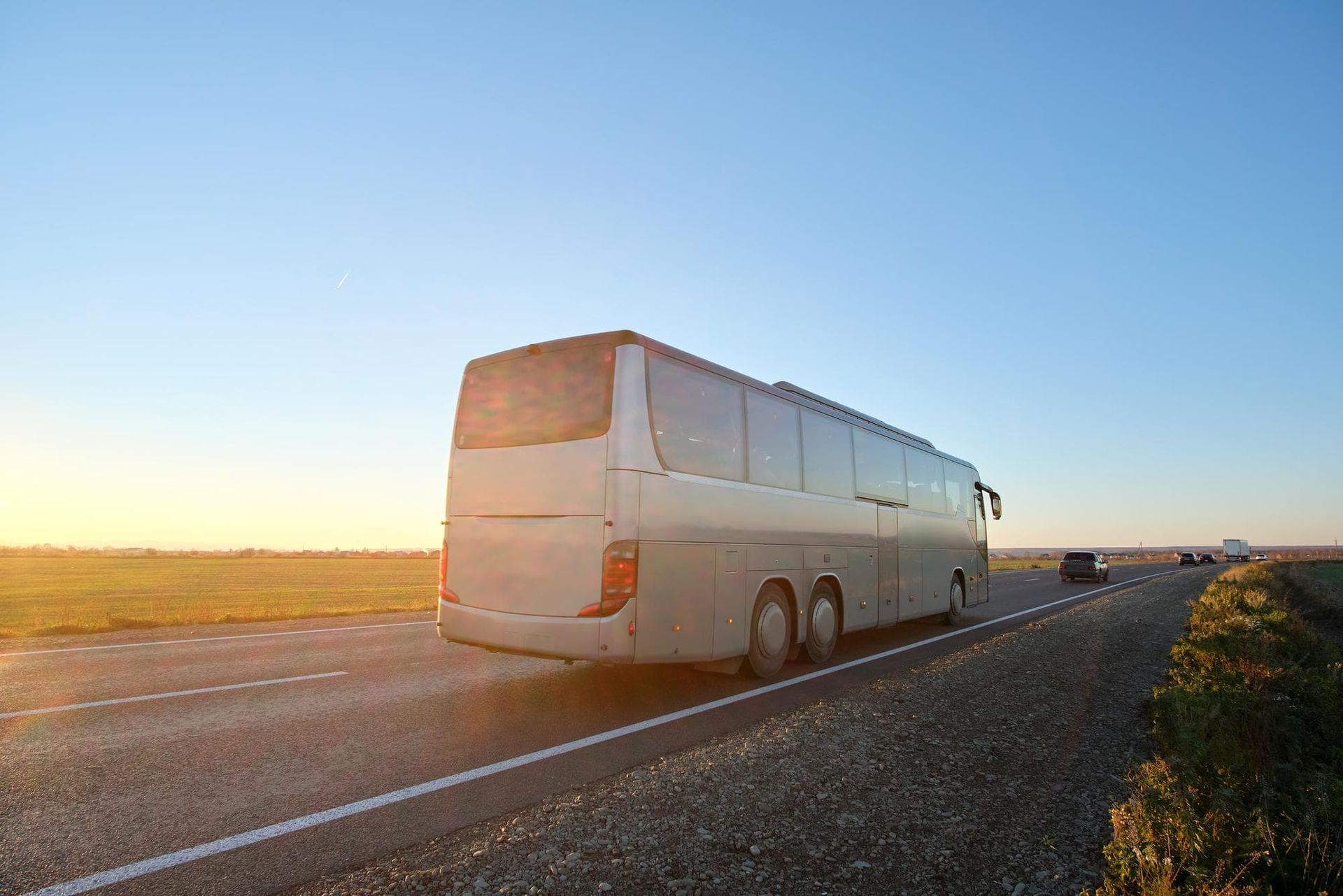 Um ônibus turístico prateado percorre uma estrada sob um céu azul, com o pôr do sol lançando um brilho quente.