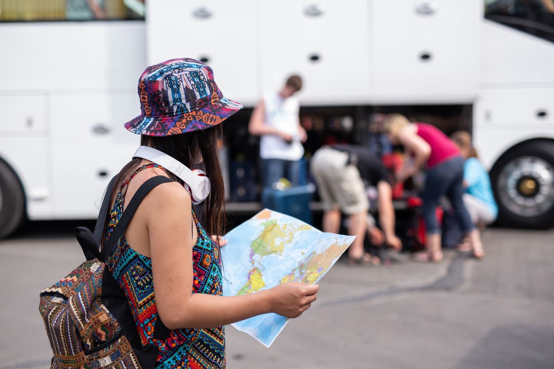Mulher de chapéu com mapa, perto de um ônibus, enquanto outras pessoas carregam suas malas.