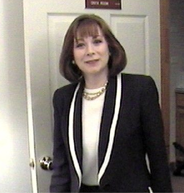 A person with short light hair and glasses wearing a black shirt with a blue and white leaf pattern against a grey circle.