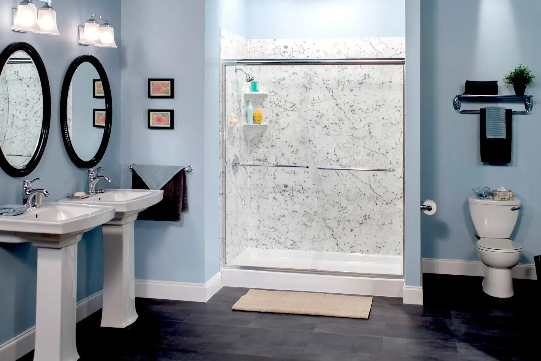 A blue-walled bathroom with two white pedestal sinks, a walk-in shower with marble-patterned walls, and a white toilet.